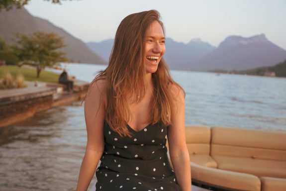 A woman in a polka dot dress smiles by a lake surrounded by mountains.