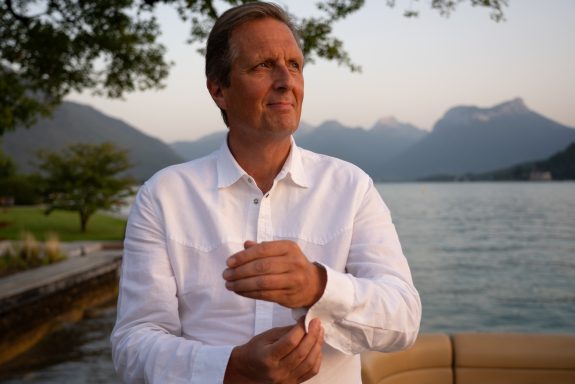 Man in a white shirt adjusting his cuff, with mountains and a lake in the background.