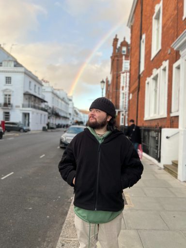 A person in a black jacket stands on a street with a rainbow in the background.