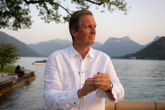 A man in a white shirt stands by a lake, mountains in the background, at sunset.